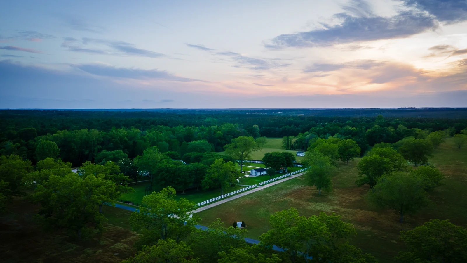 An aerial view of The Orchards at sunset, with tree-lined lawns, white fences, buildings, and countryside stretching toward the horizon.