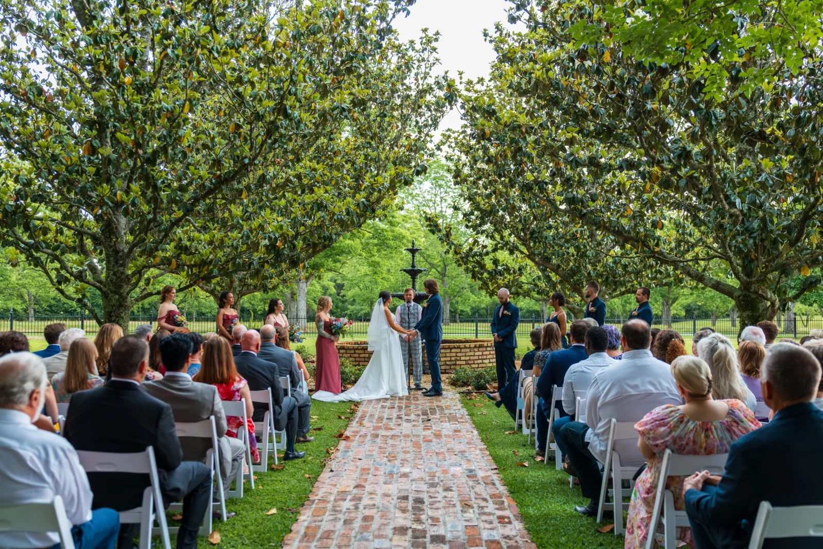 A couple exchange vows at Magnolia Fountain, surrounded by guests seated beneath the trees.