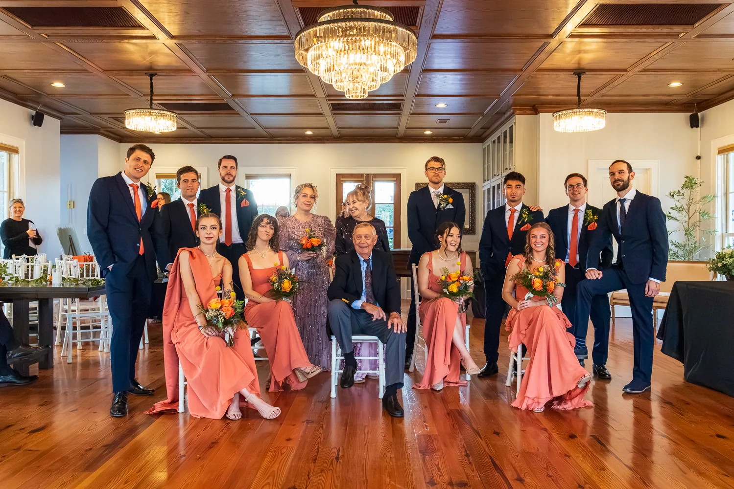 A wedding party posing for a group portrait in the Oak Room beneath crystal chandeliers and a wood-paneled ceiling.