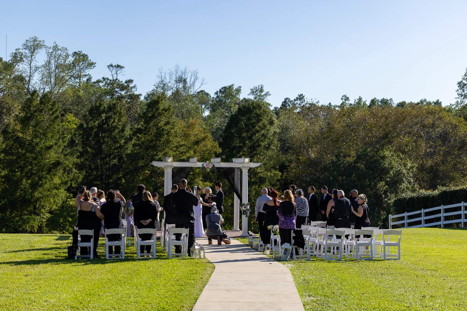 Guests standing beside rows of white chairs as a couple marry beneath the Waterfront Pergola, with the curved aisle crossing the lawn.
