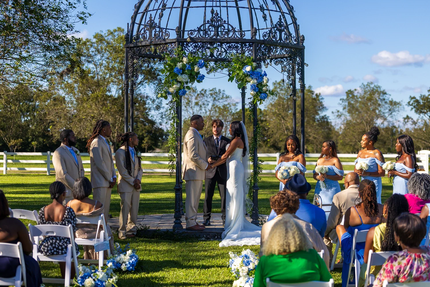 A couple exchange vows beneath the iron Rotunda decorated with blue and white flowers, surrounded by their wedding party and seated guests.
